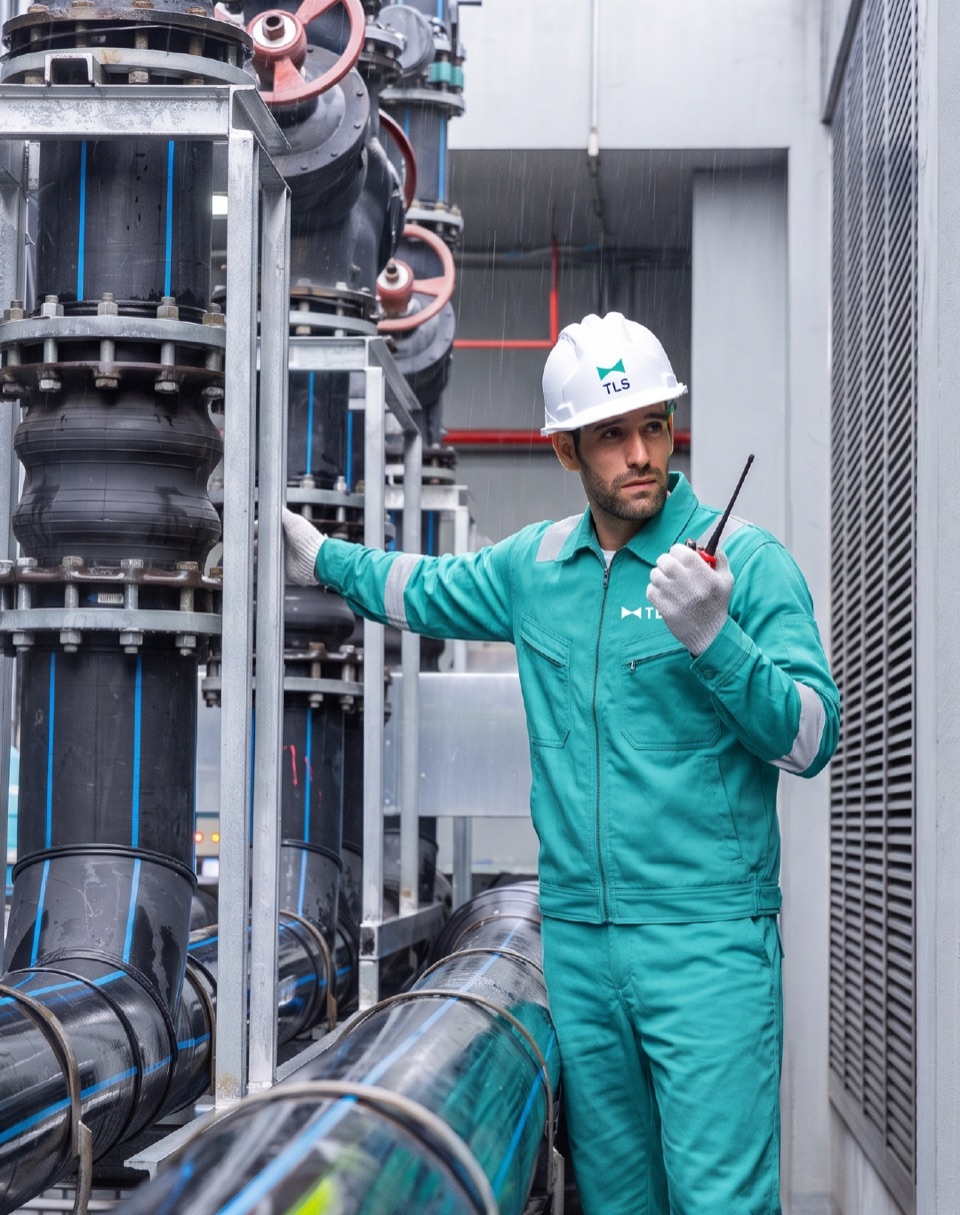 A TLS technician in a hard hat and teal coveralls checking pipework in a plant room, holding a radio